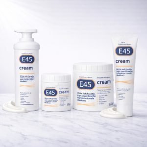 Four E45 cream products on white marble background including white pump bottle, 125g white tub, 350g white tub, and white squeeze tube with blue and orange E45 branding.