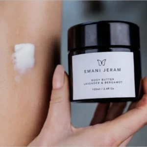 Elegant black jar of Emani Jeram Body Butter in Lemon & Eucalyptus scent, placed on a minimalist ceramic dish with soft accents of decorative dried foliage. The label features a butterfly logo, highlighting the product's luxurious and natural appeal. Contains 100ml (2.41 fl oz).